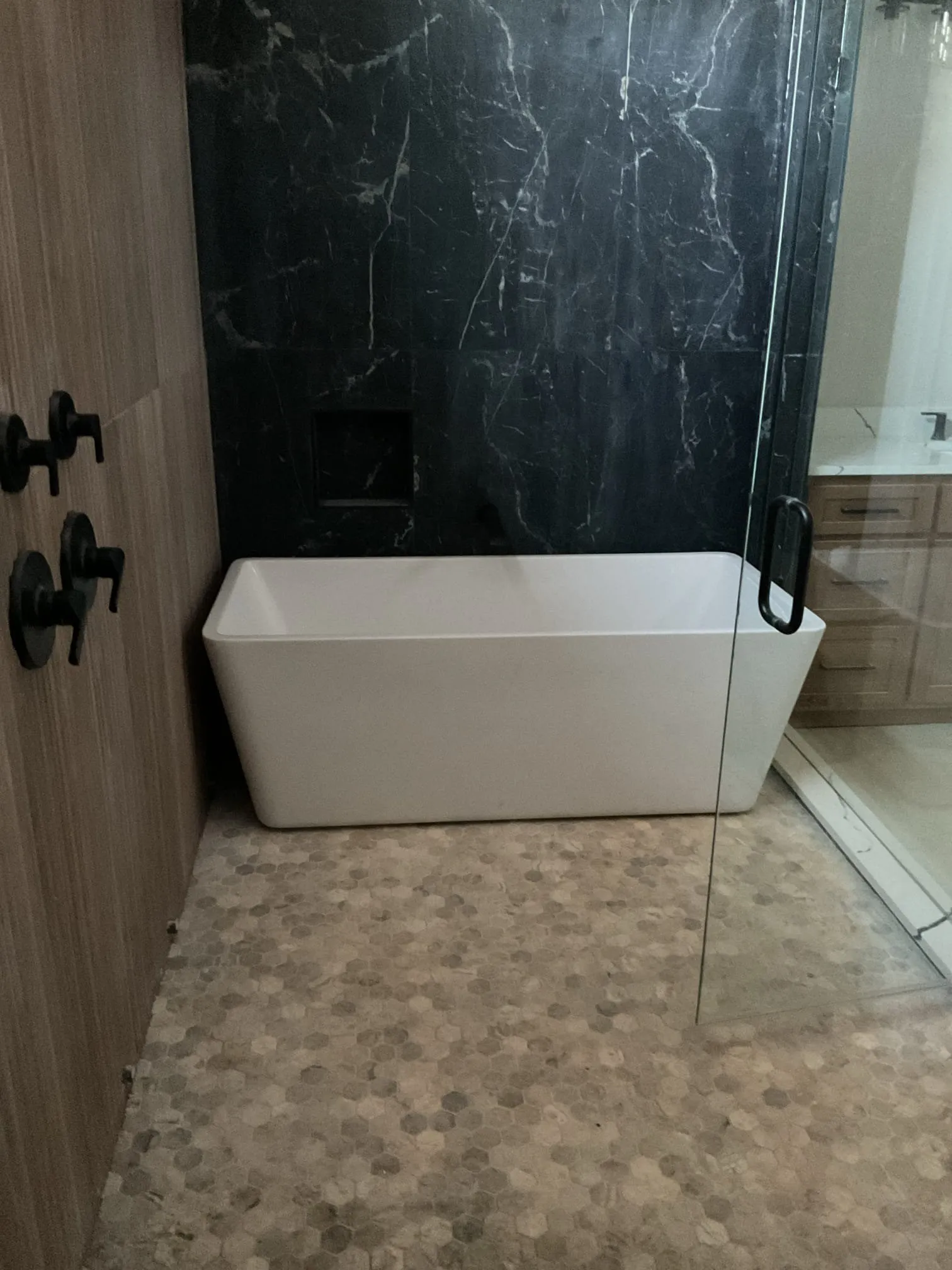 Modern bathroom with a white freestanding bathtub, black marble wall, and hexagonal tile flooring.