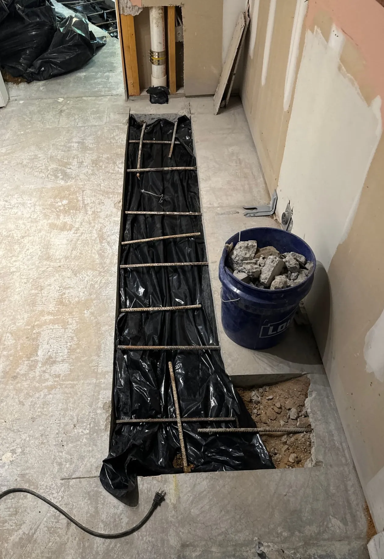 Exposed trench in a concrete floor prepared for plumbing work, with a bucket of rubble nearby.